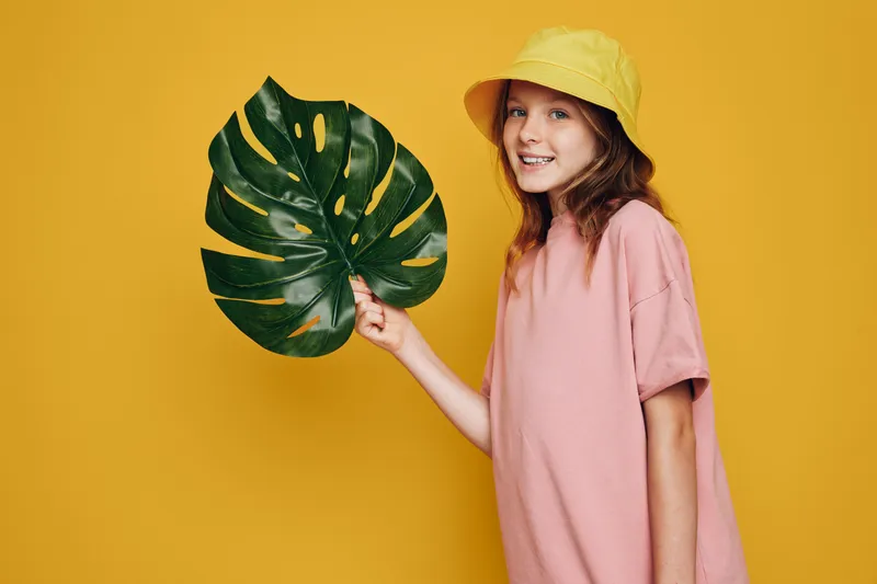 Smiling girl in a yellow hat and pink oversized t-shirt holds a large green tropical leaf against a yellow background. Bright studio portrait with summer and nature concept.