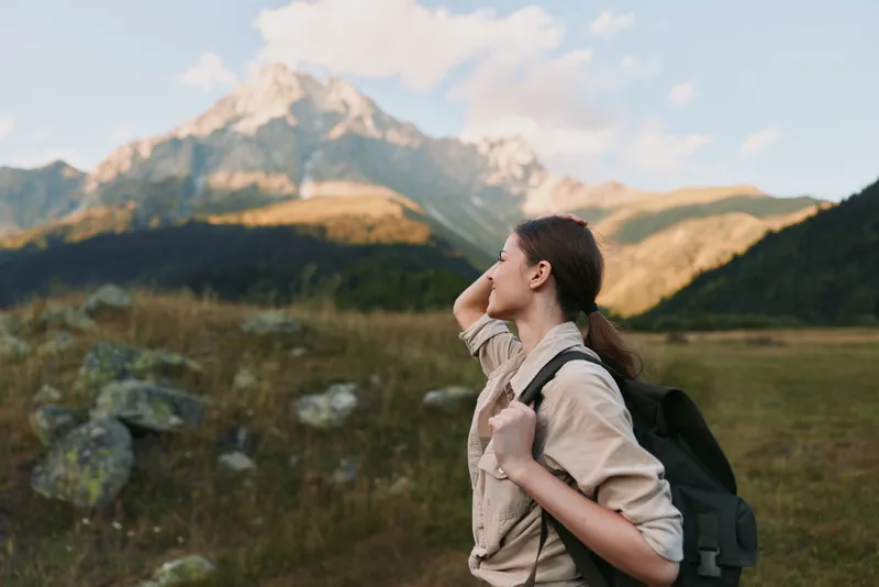 Woman hiker with backpack admires distant alpine peaks in a wide meadow, enjoying a calm outdoor lifestyle moment and fresh mountain air. She explores rugged scenery.