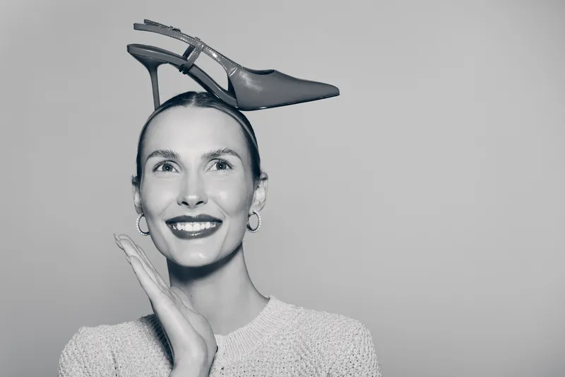 Smiling white woman with pale skin holds high heel shoe on head, wears earrings and knitted sweater. Concept for fashion, humor, and creative portrait studio shot.
