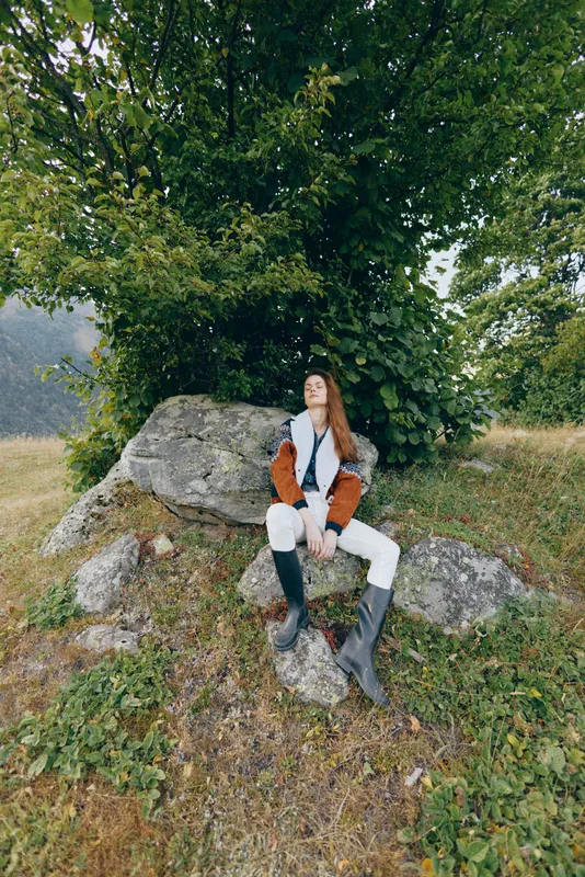 Woman in nature outdoors sitting on rocks under a large tree, relaxed and wearing boots and casual jacket, countryside portrait showing scenic grass, leafy forest and peaceful leisure moment.