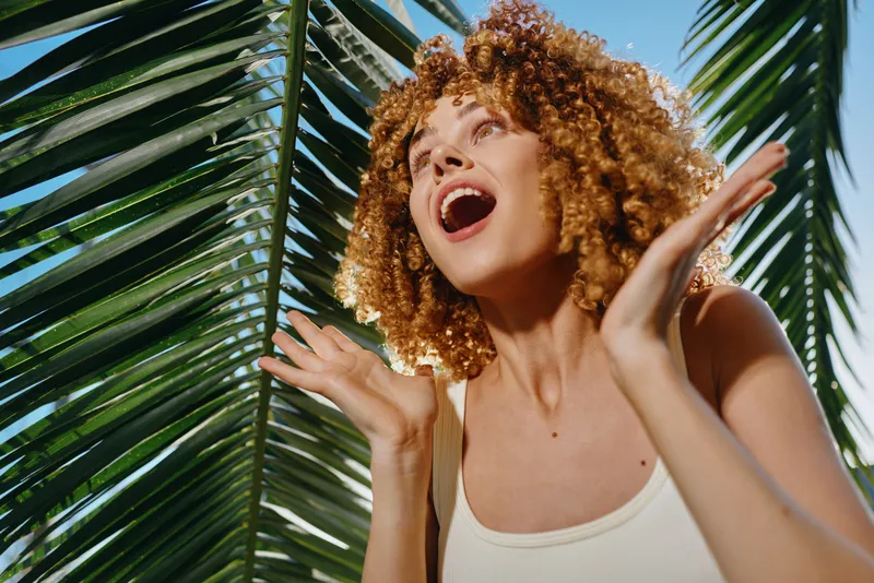 joyful woman with curly hair smiling brightly outdoors by palm leaves in warm sunlight, wearing a sleeveless top with natural tropical background.