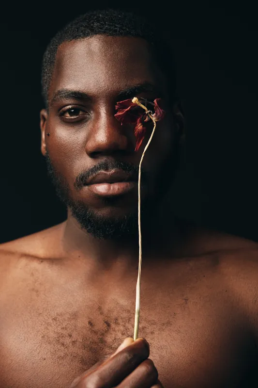 African man with dark skin holding dried flower in front of eye in creative conceptual visual style. Portrait with bare chest and serious expression on black background for artistic photography.