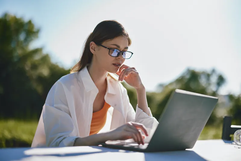 Young woman working on a laptop outdoors, wearing glasses and a casual white shirt, reflecting concentration and creativity against a bright background