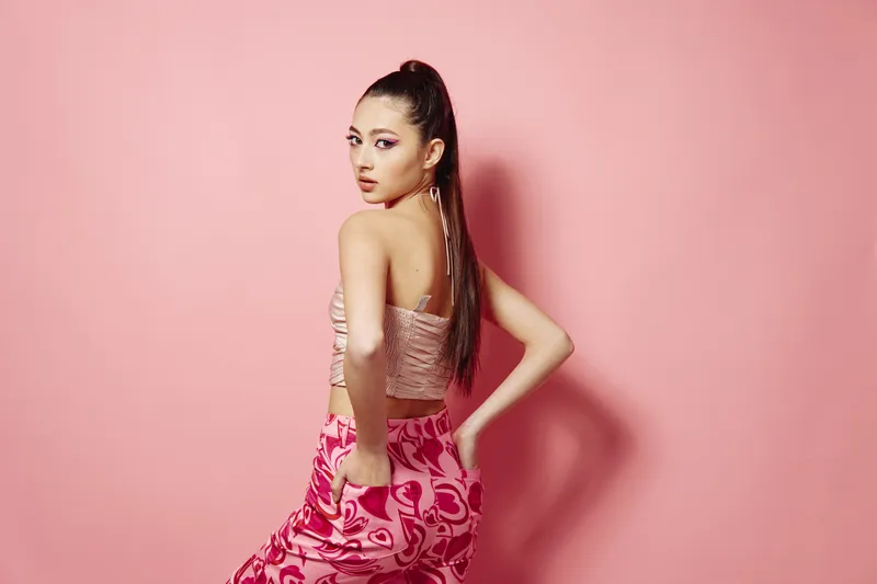 Young stylish woman in pink abstract print trousers and blush top poses against a pastel pink background. Her long hair is in a high ponytail. Concept of beauty, fashion, and creative styling.