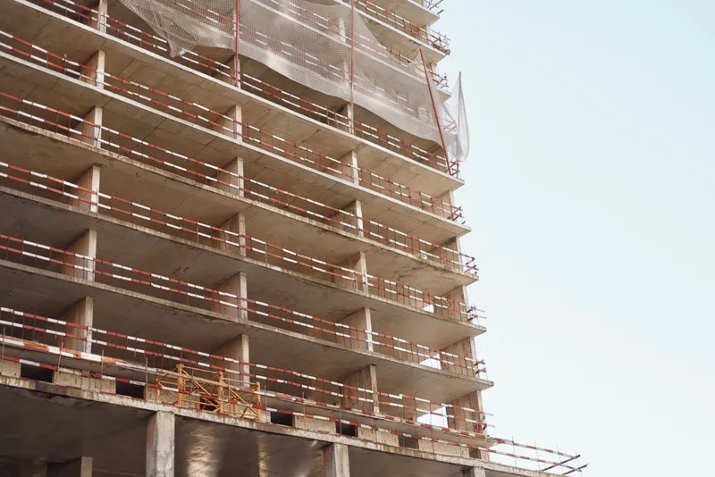 Unfinished multi-story building under construction stands against clear sky with scaffolding and safety nets surrounding concrete structure.