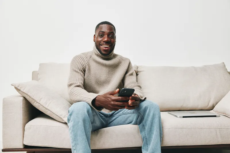 Happy young man in a cozy sweater using a smartphone on a neutral sofa, conveying comfort and modern lifestyle