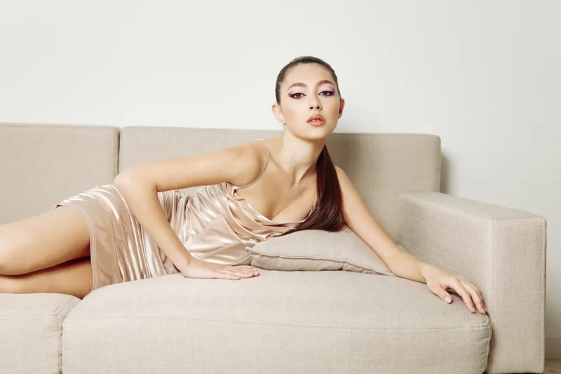 Young woman in silky beige dress lounges gracefully on a minimalist sofa with stylish makeup and sleek hair in a modern beauty fashion concept focused on elegance and creativity.