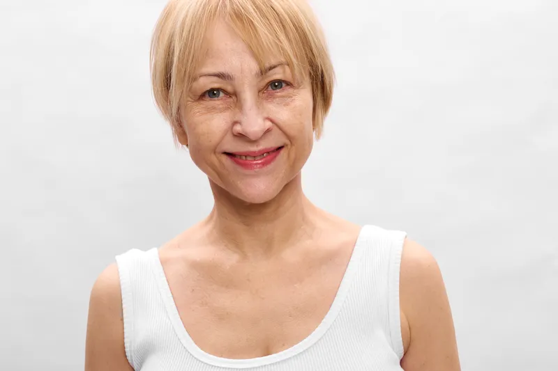 Smiling elderly woman, blond short hair, studio portrait, white top, soft light, gentle expression, skincare and wellness concept, self care on white background, calm beauty mood