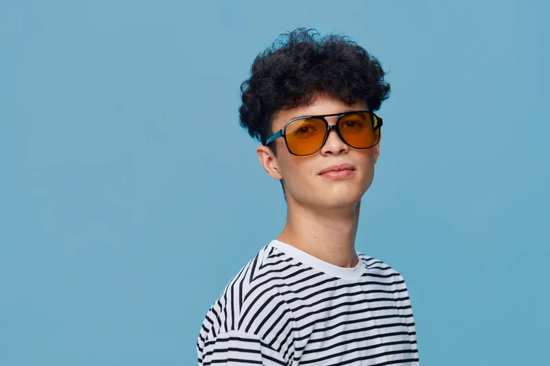 young man wearing striped shirt and oversized yellow sunglasses posing confidently against blue background with curly hair and casual style, modern portrait