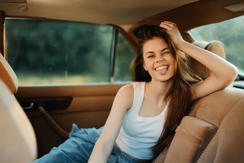 Young woman in casual attire sitting in a car, showing playful emotions with tongue out, enjoying a sunny day in a stylish environment.