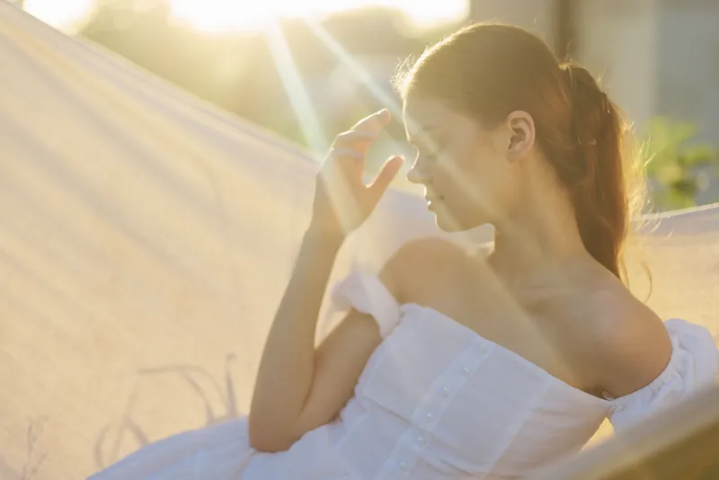 A young woman in a white dress enjoying a peaceful moment in a hammock under soft sunlight, evoking feelings of relaxation and serenity