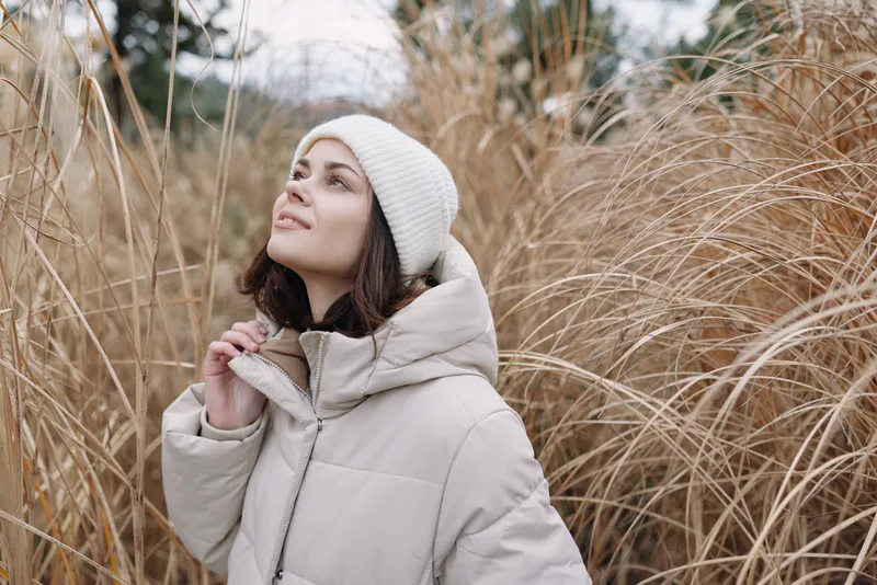 Woman in a light winter coat and white beanie gazes upward among tall grasses, capturing a serene outdoor portrait in soft natural light.