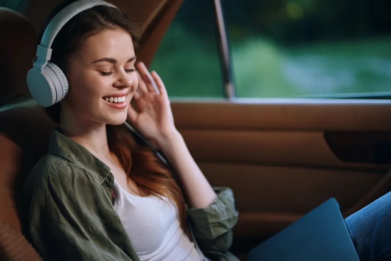 Young woman enjoying music with headphones while relaxing in a car, expressing happiness and tranquility in a natural setting.