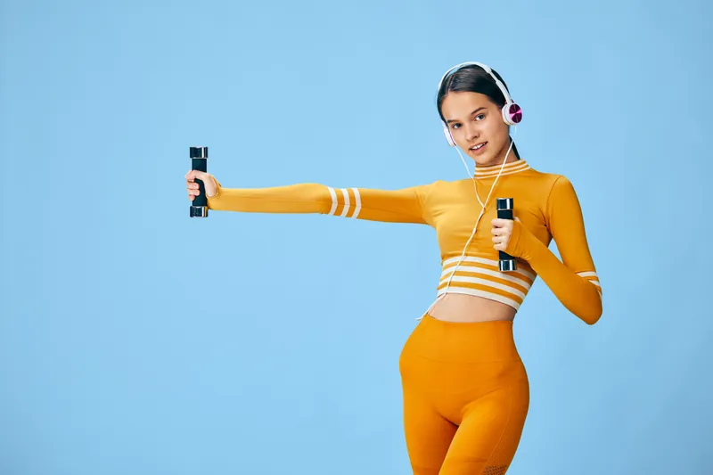 Fit woman in orange activewear exercising with dumbbells and wearing headphones against a plain light blue studio background, showcasing fitness, strength and an energetic healthy lifestyle.