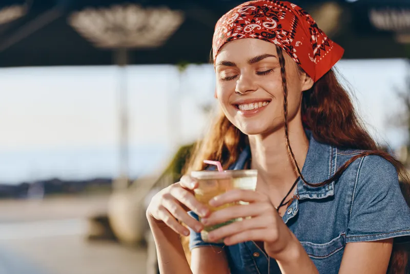 smiling woman enjoying refreshing drink outdoors with casual denim outfit and red bandana in warm sunlight during summer day