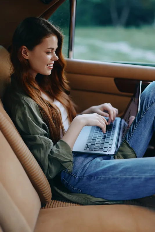 Young woman in casual attire working on a laptop inside a car, showcasing remote work, comfort, and a relaxed lifestyle, with natural lighting and green background.