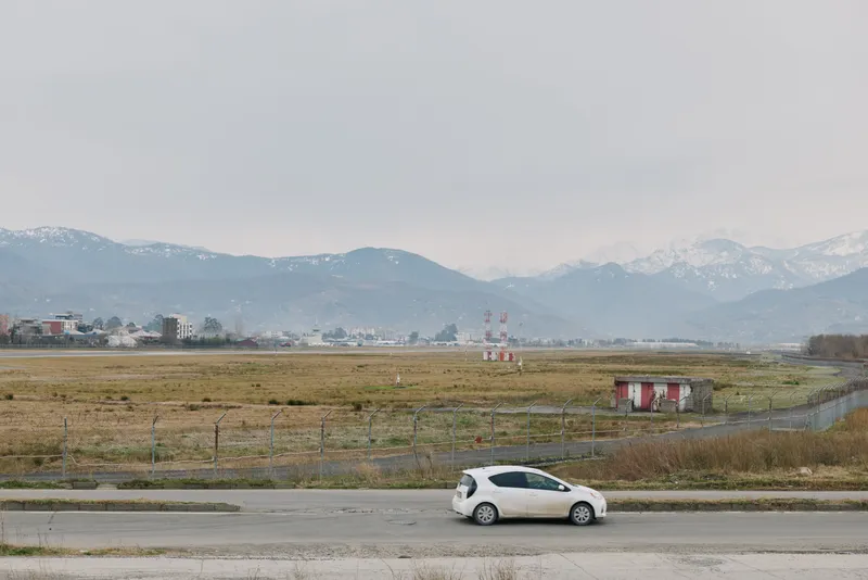 White compact car parked on roadside near field with distant mountains under cloudy sky. Concept of travel, transportation, countryside, and nature landscape in calm weather conditions.
