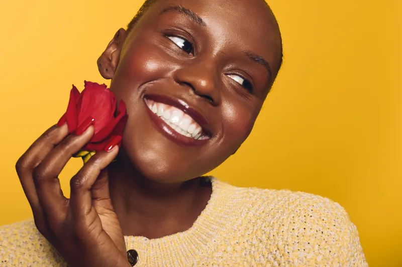 Happy African woman with dark skin holding red rose near face, smiling and looking away. Bright yellow background and cozy sweater create warm, joyful mood. Concept of beauty and happiness.