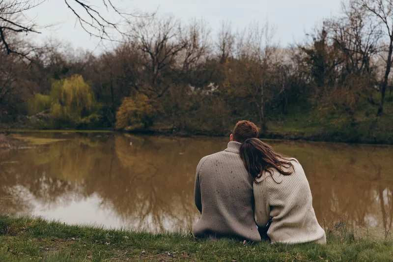 Couple sits by a lakeside in autumn, cozy sweater, view of water, tranquility and friendship in a serene outdoor moment, nature scene with soft light, calm colors, relaxing companionship