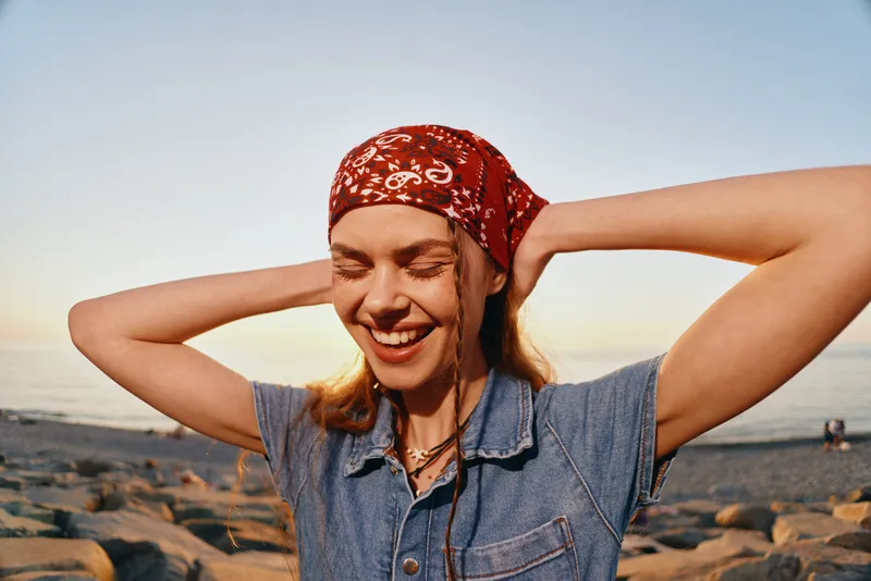 Lifestyle woman in modern boho-western denim street style smiling outdoors at sunset with film color effect and joyful expression near rocky beach shore.