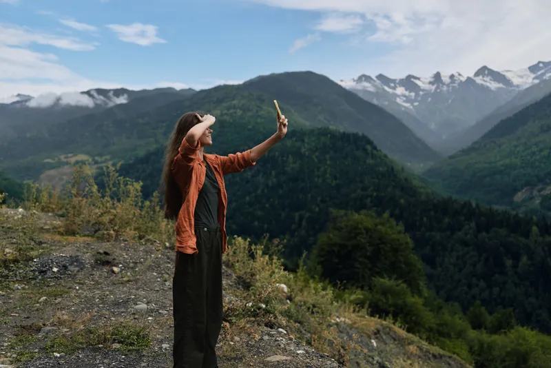 Outdoor scene with a woman taking a selfie on a rocky overlook in a mountainous landscape, capturing a vivid moment amid green valleys and blue skies.