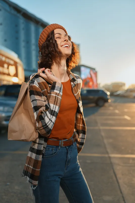 Woman shopping portrait smiling in urban street at golden hour, candid authenticity and lifestyle moment with beanie and paperbag, mindful living and emotional storytelling in city glow