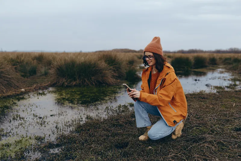 Woman lifestyle outdoors, kneeling by a river and grassy field, casual orange jacket and hat, using a smartphone, serene nature mood in autumn overcast daylight