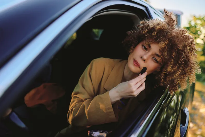 Cheerful woman in a car applying makeup, sunlit outdoors, retro film color, relaxed pose, candid moment, natural tones, warm atmosphere, greenery in the background, fresh breeze and sunlit