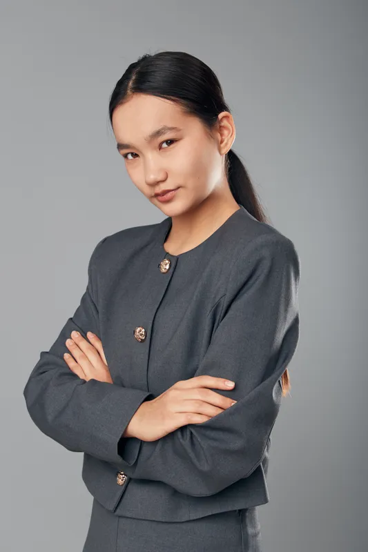Woman with crossed arms wearing formal jacket standing in studio with neutral gray background looking confidently at camera, professional young businessperson portrait with neat hairstyle.