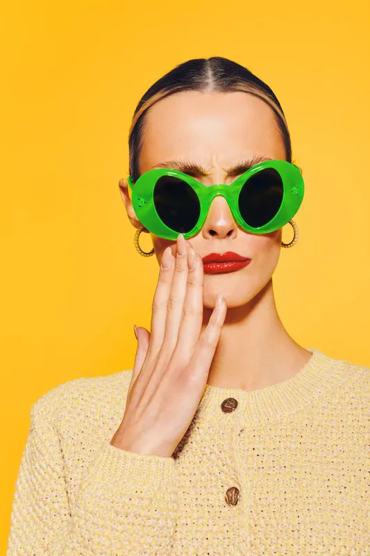 Young woman with green sunglasses and red lipstick touches her face with a worried expression. Bright yellow background and beige sweater create a vivid contrast. Concept of concern.