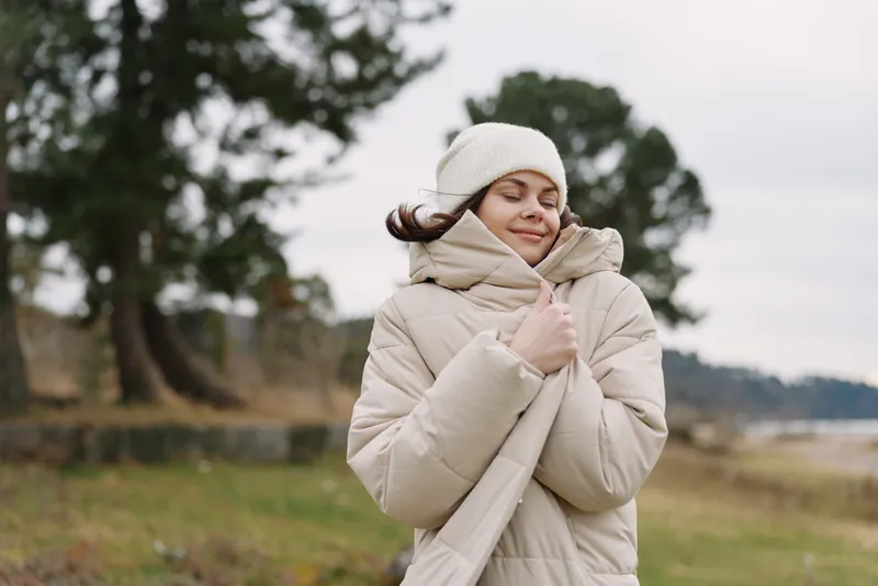 Winter scene with a woman in a beige puffer coat and beanie, wrapped in a scarf, enjoying a windy outdoor landscape and a calm, cozy mood.