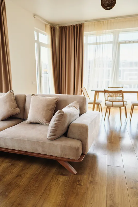 Sofa in a bright living area with cushions, wooden floor and dining setup by large windows, neutral curtains, minimalist modern interior flooded with warm natural light.