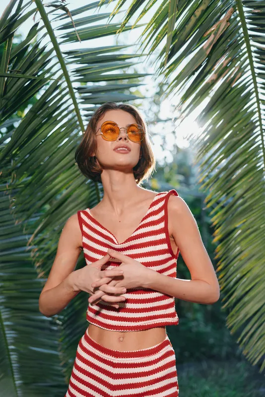 A stylish woman stands beneath tropical palm leaves wearing a red and white striped crop top and matching shorts. She wears orange sunglasses and exudes relaxed summer confidence with a sunny,
