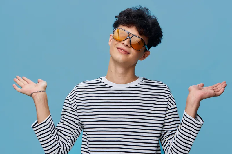 young man shrugging shoulders with playful expression wearing orange sunglasses and striped shirt against blue background, casual style, positive mood
