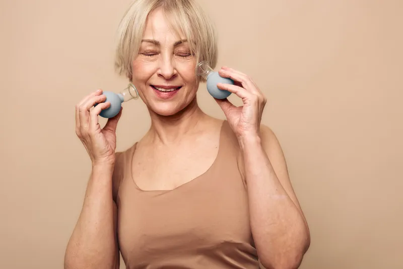 Happy middle aged woman with closed eyes using facial massage rollers on cheeks. Skin care and anti aging concept with natural beauty and wellness on beige background in soft light.