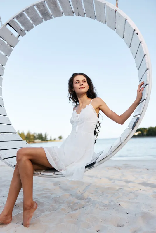 A young woman sits gracefully on a circular wooden frame on a sunlit beach, wearing a white dress, exuding calm elegance and a breezy, carefree summer mood.