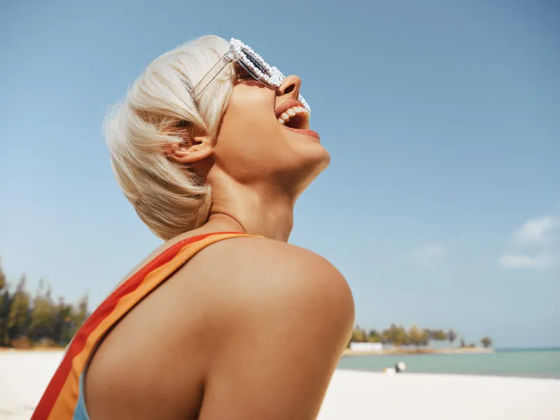 Joyful sunlit portrait of a smiling woman at the beach, wearing sunglasses, with head tilted back and laughter echoing. Warm glow, salty breeze, vivid color, and carefree summer mood.