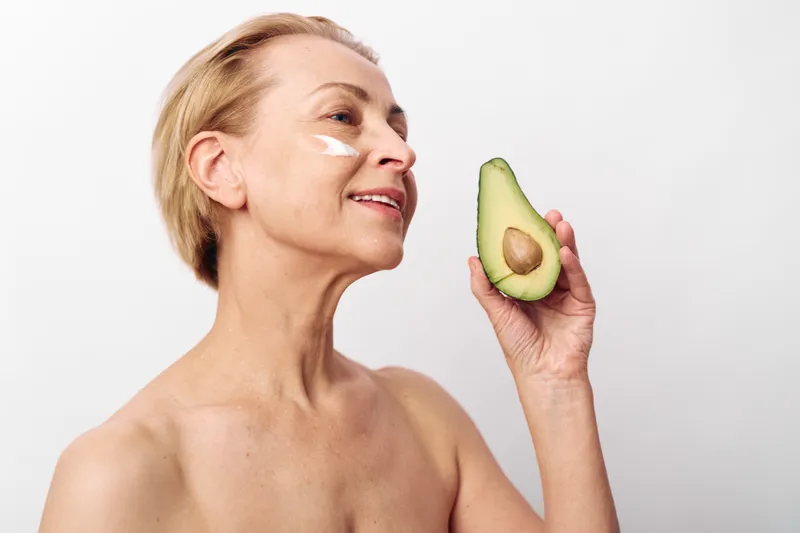 Mature woman with natural skin holds avocado and applies white cream on cheek, smiling softly. Concept of skincare, organic beauty, healthy lifestyle, and natural facial care at home.