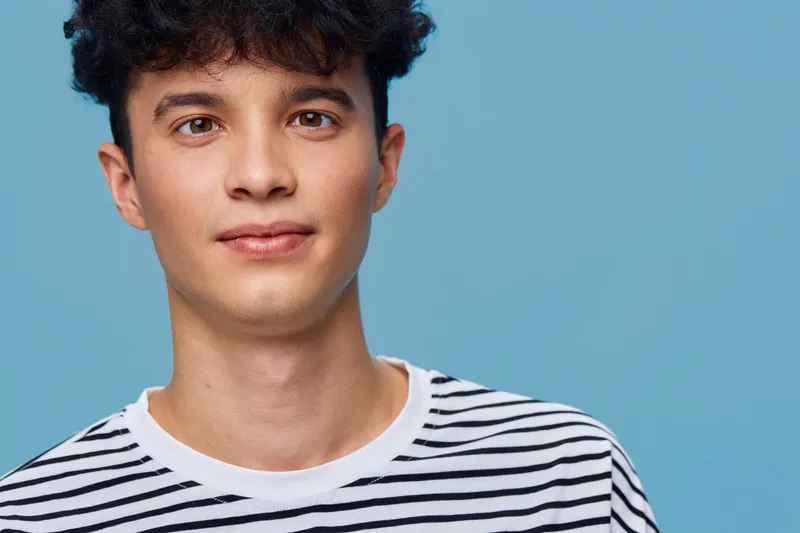 Young man with curly hair wearing a striped shirt poses against a solid light blue background, showcasing a relaxed and casual expression.