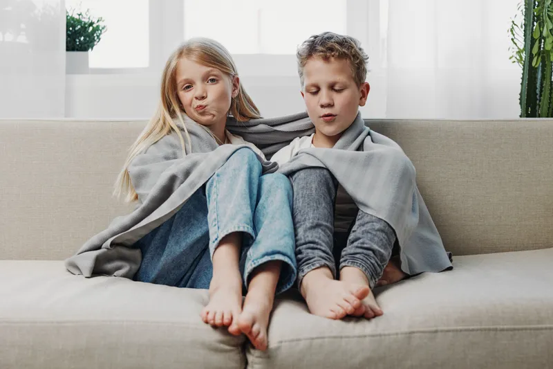Two children wrapped in a blanket sit barefoot on a sofa near a bright window, showing relaxed and cozy mood at home.