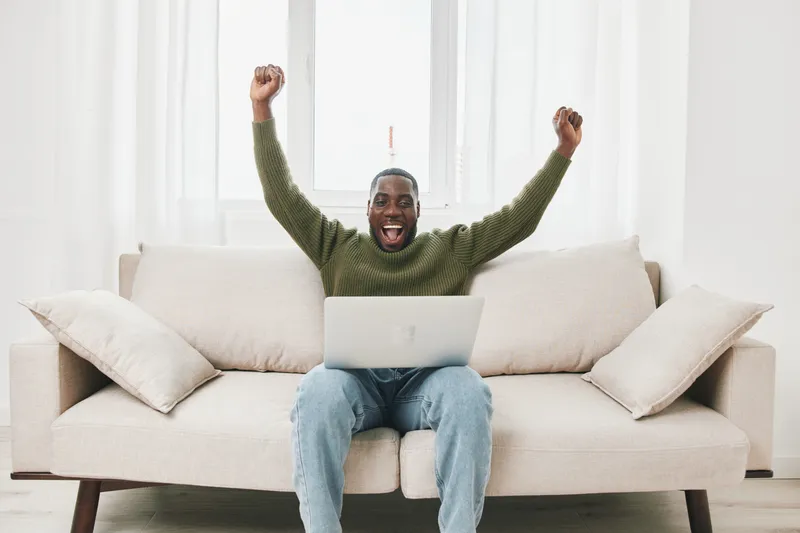 Joyful man celebrating success while using a laptop in a bright living room setting, wearing a cozy sweater, showcasing happiness and accomplishment