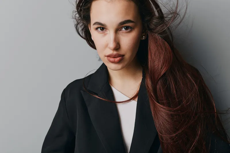 Portrait of confident woman with long hair wearing black coat and white top against gray background. Female model looking at camera with natural makeup and serious expression.