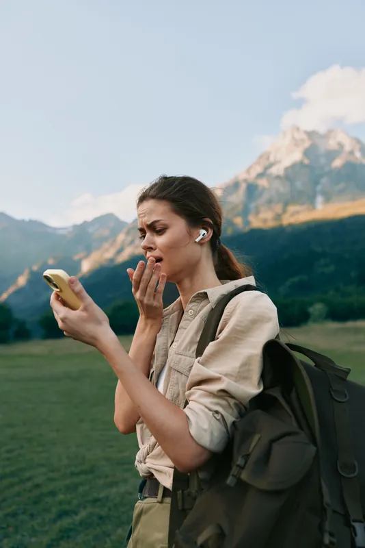 A woman outdoors with a backpack checks her phone, standing in a sunlit meadow with mountains behind. A candid lifestyle moment focused on travel, exploration, and curiosity.