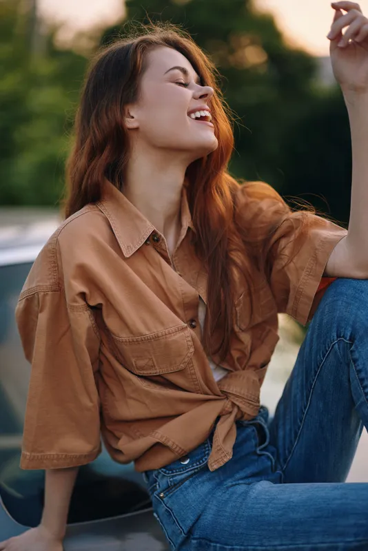 Young woman with long hair wearing a stylish brown shirt and blue jeans, enjoying the outdoors with a cheerful expression during sunset.
