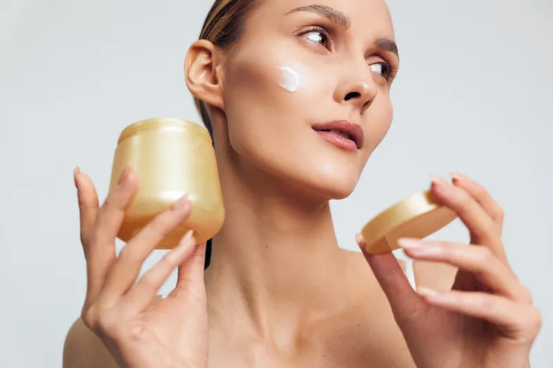 A woman applies skincare cream on her cheek, holding two gold jars, presenting a simple beauty routine for glowing skin, isolated on a clean studio background.