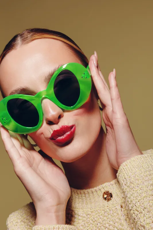 Young woman with bright red lipstick wearing large green sunglasses and beige sweater poses against neutral background. Concept of fashion, style, and summer accessories.