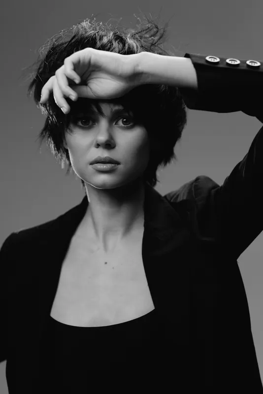 Black and white portrait of a young woman with short hair shielding her eyes, dressed in dark clothing, expressing curiosity and intrigue.