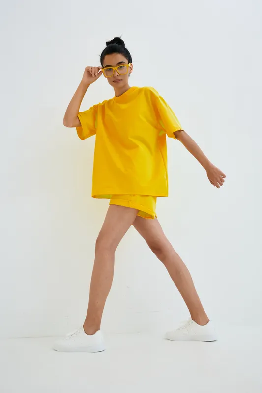 Young woman in bright yellow casual outfit walking confidently indoors, wearing matching shorts, oversized t-shirt, and stylish glasses on a plain white background.