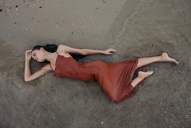Solitary woman lies on sandy ground in a flowing red dress, creating a dramatic, artistic pose with soft natural light and muted textures across the surface.