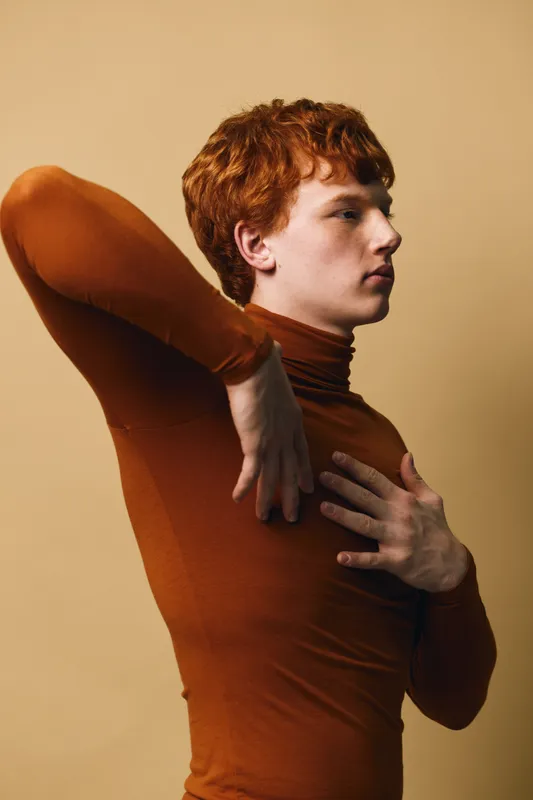 Young man with red hair wearing brown turtleneck sweater posing in profile with hand on chest and arm raised against beige background in studio.